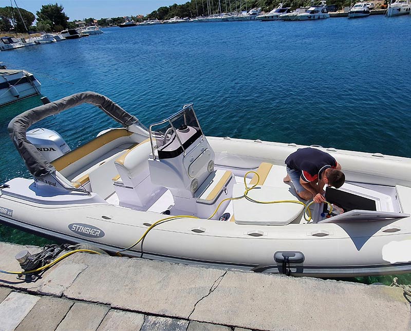 Man kneeling on a white inflatable boat at a concrete dock, inspecting an open hatch by a yellow hose in a sunny marina.